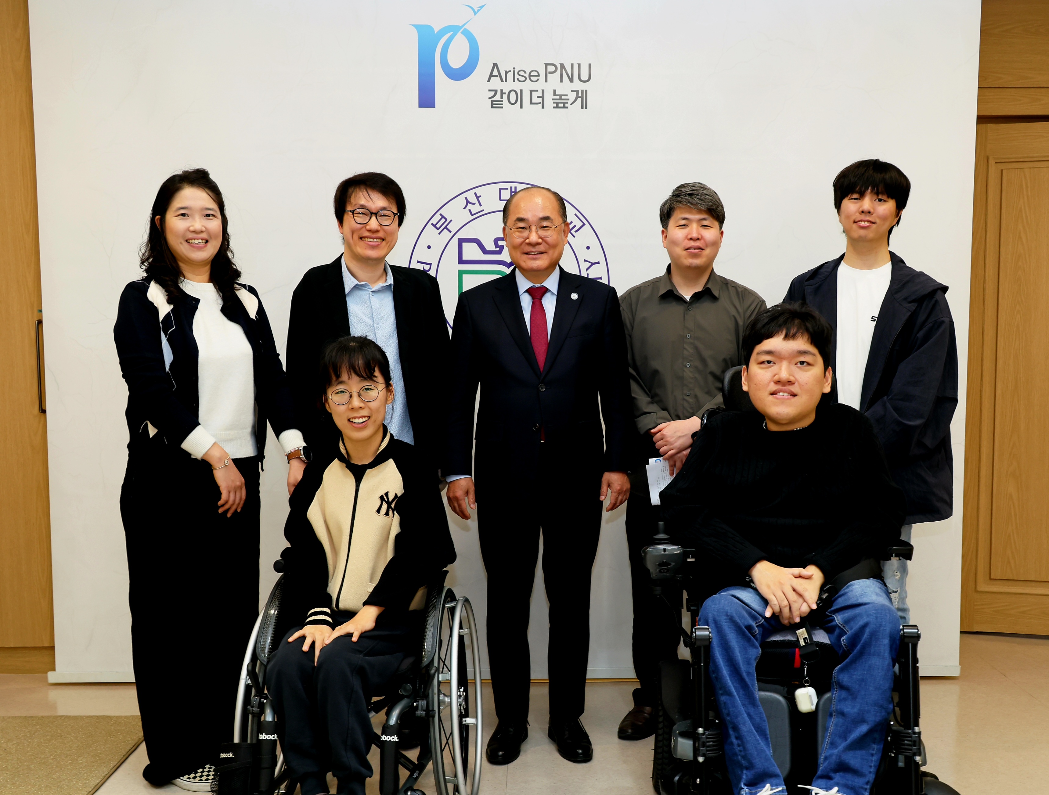 부산대학교 로고를 배경으로 찍은 단체사진. 가운데 부산대학교 총장을 중심으로 장애학생지원센터 관계자와 장애대학원생이 둘러싸고 있다.