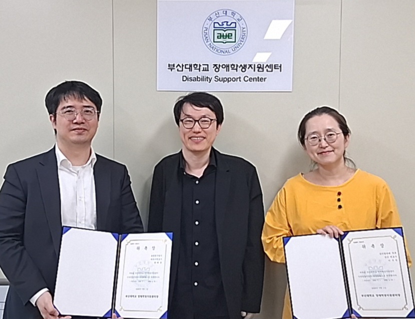 부산대학교 장애학생지원센터 현판 아래에서 임용재 장애학생지원센터장을 중심으로 권태균 변호사(로엘법무법인)와 이주언 변호사(공익법단체 두루)가 위촉장을 들고 서 있다.